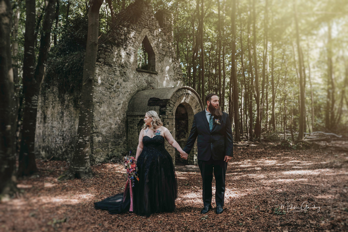 A couple holding hands before the woodland chapel ruins at Killyon Manor